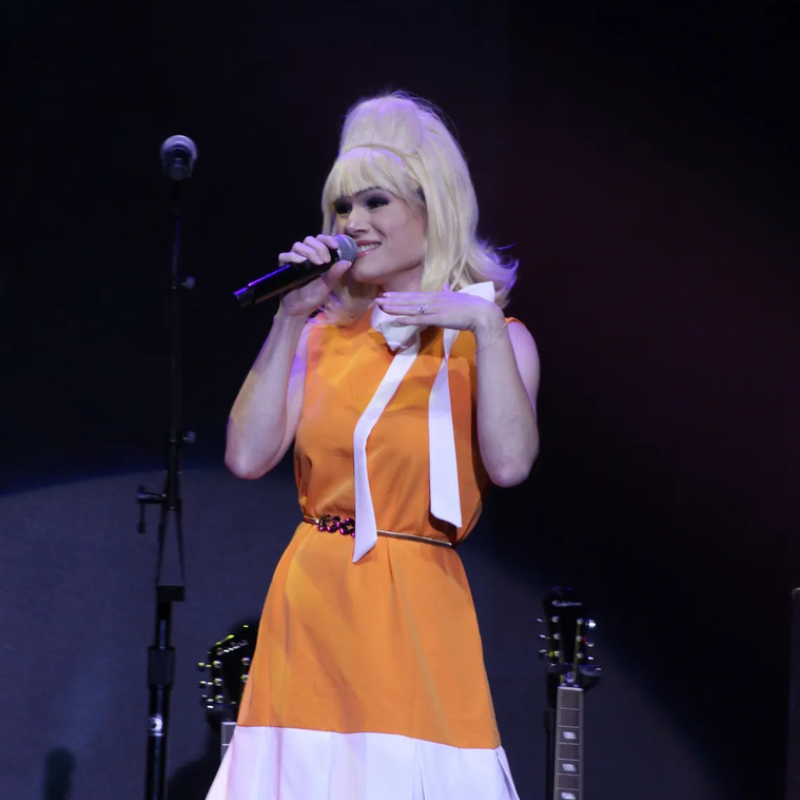 Sixtiesmania® 60s tribute band female vocalist singing live on stage in an orange 1960s-inspired dress with a blonde beehive hairstyle.