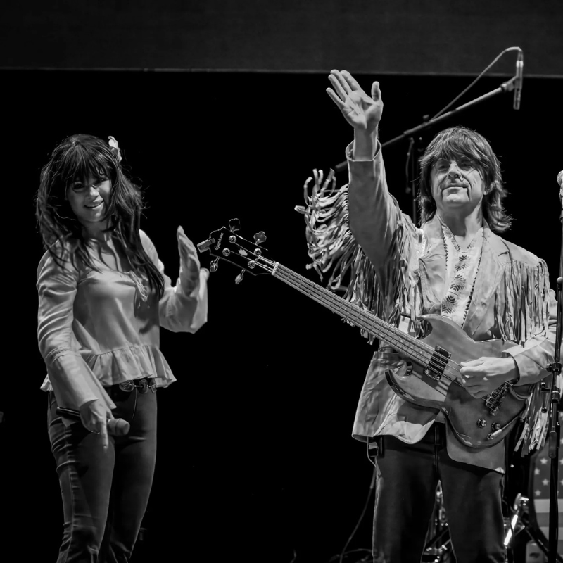 Sixtiesmania® 60s tribute band bassist and female vocalist waving to the audience during a live performance in 1960s-inspired costumes.