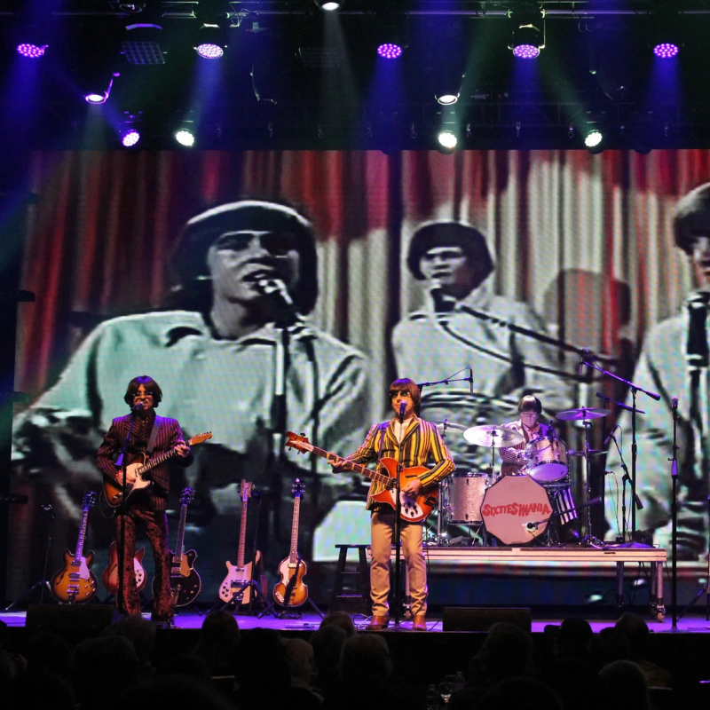 Sixtiesmania® 60s tribute band performing live with electric guitars and drums in front of a large screen featuring vintage 1960s music footage.
