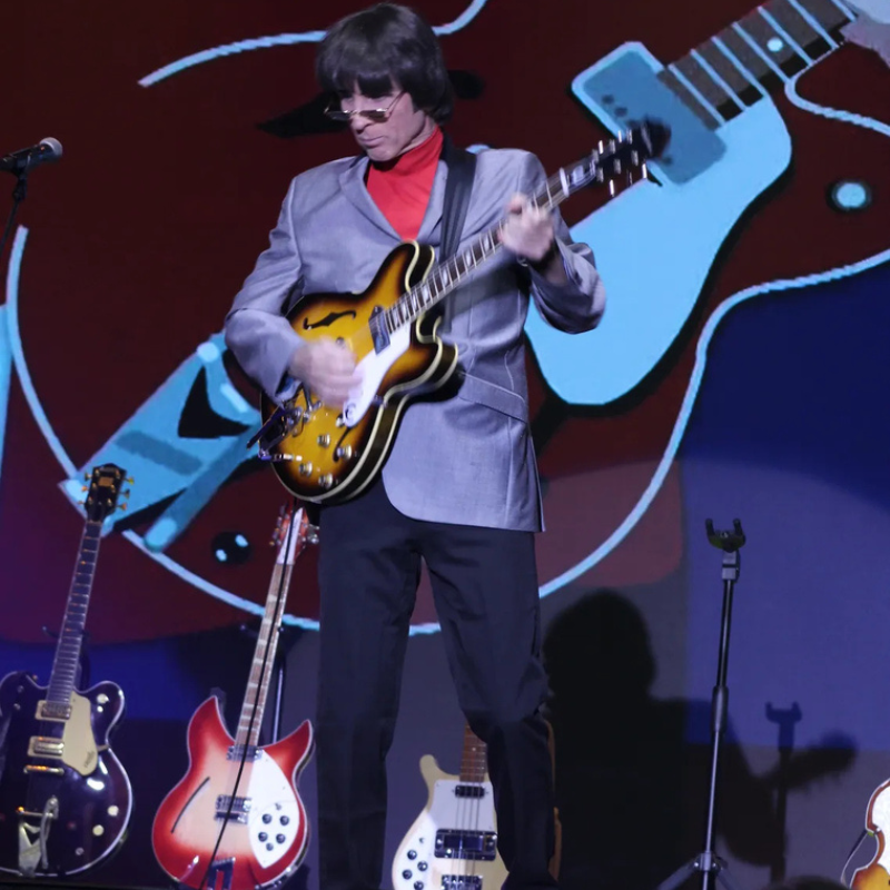Sixtiesmania® 60s tribute band guitarist playing a sunburst electric guitar on stage in a gray jacket and red turtleneck during a live Beatles tribute performance.