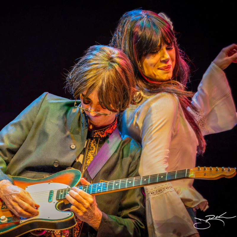 Sixtiesmania® 60s tribute band guitarist playing an electric guitar alongside a female vocalist in vintage 1960s-inspired attire during a colorful live performance.